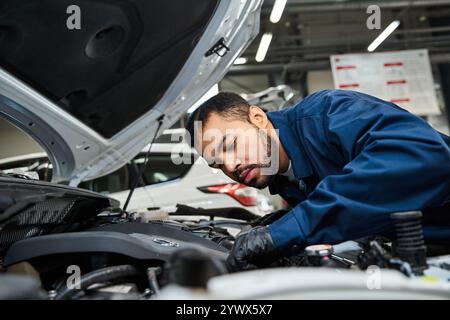 Beau mécanicien répare un moteur de voiture tout en se concentrant intensément dans un espace d'atelier occupé. Banque D'Images