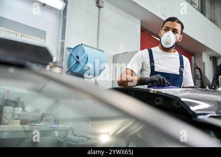 Jeune mécanicien en salopette se concentre sur sa tâche tout en portant un masque et des gants dans un atelier Banque D'Images