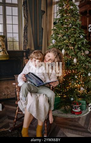 Temps de lecture intérieur confortable par le sapin de Noël avec enfant pendant la saison des fêtes Banque D'Images