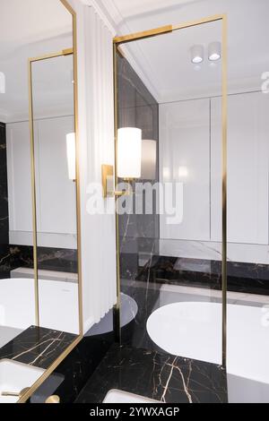 Une salle de bains avec des carreaux de métro verticaux en marbre et empilés, un meuble-lavabo en chêne blanc, des miroirs carrés encadrés de noir et des robinets, et un comptoir en marbre. Banque D'Images
