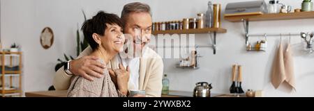 Un couple heureux partage une étreinte joyeuse dans leur charmante cuisine remplie de lumière chaude. Banque D'Images