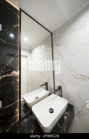 Une salle de bains avec des carreaux de métro verticaux en marbre et empilés, un meuble-lavabo en chêne blanc, des miroirs carrés encadrés de noir et des robinets, et un comptoir en marbre. Banque D'Images