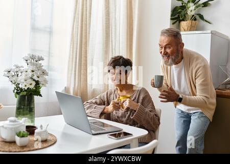 Couple mature apprécie la compagnie des autres tout en naviguant sur leur ordinateur portable dans une cuisine ensoleillée. Banque D'Images