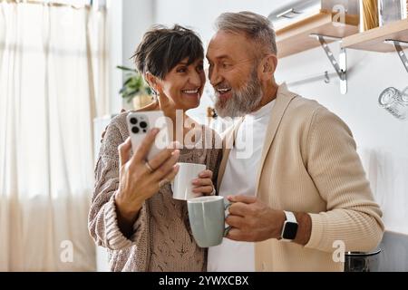 Couple senior partage un moment joyeux, tenant des tasses et capturant des souvenirs dans une cuisine ensoleillée. Banque D'Images