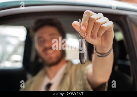 Heureux jeune homme montrant les clés de voiture à l'intérieur du véhicule neuf. Conducteur masculin joyeux célébrant l'achat de voiture ou le test de conduite réussi. Mise au point sélective sur la main h Banque D'Images