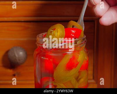 Gros plan d'une main à l'aide d'une fourchette pour soulever et égoutter quelques tranches de piments jalapeno rouges et verts, marinés dans du vinaigre d'alcool, d'un bocal en verre. Banque D'Images