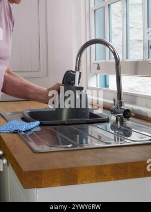 Gros plan de la main d'un homme âgé tenant une bouilloire sous l'eau courante d'un robinet au-dessus d'un évier de cuisine près d'une fenêtre à guillotine partiellement ouverte. Banque D'Images