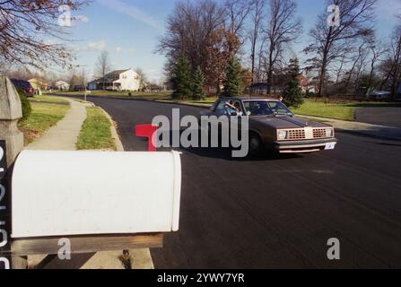 PA, USA, approx. 1985. Homme livrant des journaux dans un quartier, en les jetant dans la cour depuis la fenêtre d'une voiture. Banque D'Images