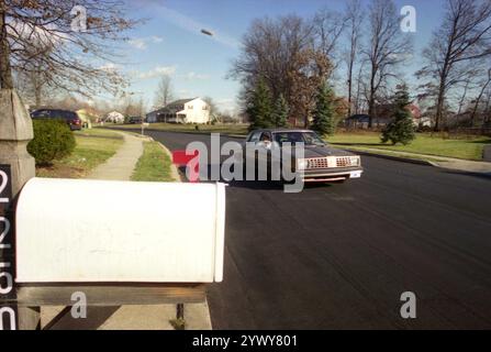 PA, USA, approx. 1985. Homme livrant des journaux dans un quartier, en les jetant dans la cour depuis la fenêtre d'une voiture. Banque D'Images