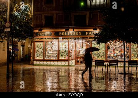 12 décembre 2024, Barcelone, Espagne : dans le quartier gothique de Barcelone, les lumières ​​the d'un magasin se reflètent sur le sol tandis qu'un homme marche pendant une soirée pluvieuse. Crédit : Jordi Boixareu/Alamy Life News Banque D'Images