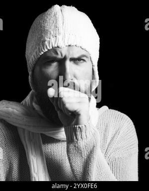 Gros plan d'un malade barbu toussant dans son poing. Santé et médecine. Malade homme malade en pull tricoté, écharpe et chapeau ayant mal à la gorge Banque D'Images