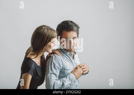 Une jeune femme avec un tatouage sur son bras gauche murmure quelque chose à un jeune homme dans une chemise bleu clair, les deux debout sur un fond blanc Uni Banque D'Images