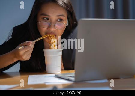 Femme d'affaires asiatique mangeant des nouilles de tasse instantanée tout en travaillant sur ordinateur portable au bureau tard dans la nuit. Focales sélectives. Banque D'Images