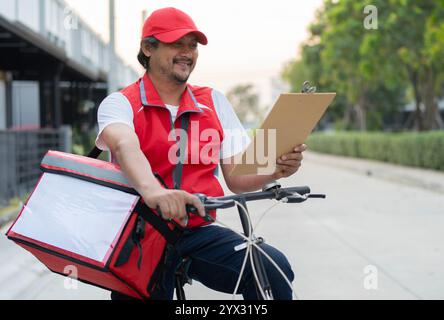 Vélo, livraison heureuse homme asiatique vérifiant le papier pour les services, fintech et emplacement de commerce électronique. Chauffeur logistique avec sac pour une commande en ligne rapide Banque D'Images