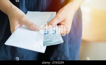 Mains tenant l'enveloppe avec de l'argent thaïlandais et sort l'argent d'une enveloppe, gros plan. Corruption de pot-de-vin, salaire, gain et concept d'épargne Banque D'Images