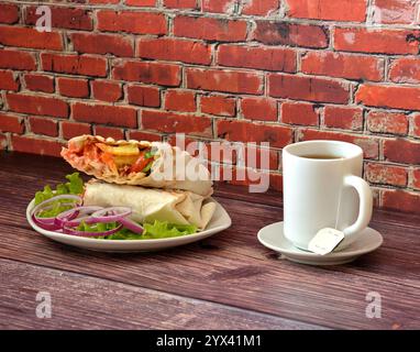 Une assiette avec shawarma de viande juteuse sur une feuille de laitue avec oignon rouge et une tasse de thé noir ensaché sur une table en bois. Gros plan. Banque D'Images