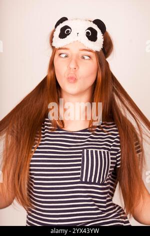 Souriant jeune femme rousse heureuse portant un masque de sommeil rayé se sentant fraîche après une bonne nuit de sommeil sain le matin isolé Banque D'Images