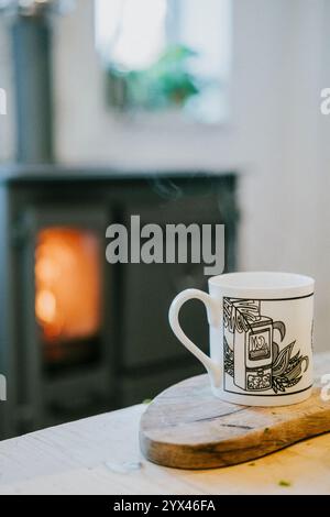 Une tasse blanche avec un motif noir se trouve sur une planche à découper en bois à côté d'un poêle à bois. La scène transmet une atmosphère cosy et chaleureuse, avec le poêle à bois Banque D'Images