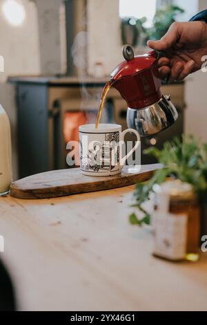Une personne verse du café dans une tasse à partir d'une cafetière rouge. La cafetière est sur une table en bois à côté d'un pot de miel. La scène transmet un et confortable Banque D'Images