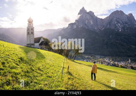 Femme en imperméable jaune profitant du coucher de soleil au-dessus des montagnes des Dolomites et de la vieille église en pierre dans le Tyrol du Sud, Italie. Banque D'Images