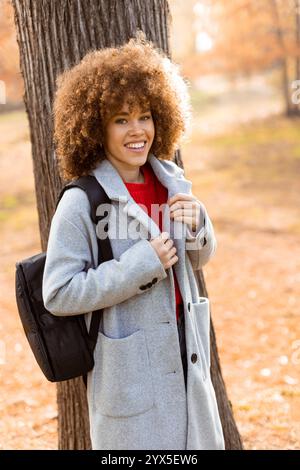 Une jeune femme joyeuse se tient près d'un grand arbre, vêtue d'un élégant manteau gris et d'un haut rouge, avec les couleurs vibrantes des feuilles d'automne qui renforcent son ch Banque D'Images