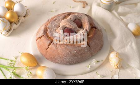 Pain de Pâques avec deux oeufs rouges nichés dans la pâte, entouré d'oeufs peints dorés et blancs, et de délicates fleurs blanches sur fond clair Banque D'Images