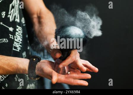Les mains des hommes tiennent la brosse à raser à partir de laquelle le talc se disperse. Styliste masculin avec des outils de rasage sur un fond noir. Coiffeur au travail. Soin des cheveux et de la barbe. Banque D'Images