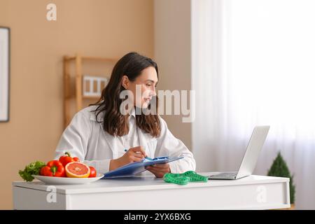 Jeune nutritionniste féminine avec presse-papiers et ordinateur portable travaillant à table dans le bureau Banque D'Images
