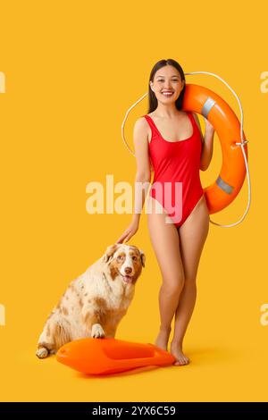 Jeune femme en maillot de bain tenant l'anneau de bouée de sauvetage et caressant chien berger australien sur fond jaune Banque D'Images