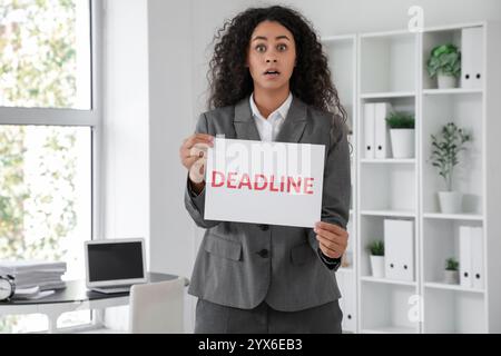 Femme d'affaires afro-américaine choquée tenant le papier avec le mot DÉLAI dans le bureau Banque D'Images