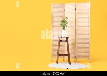 Table avec tiges de bambou dans vase, tapis et écran pliant sur fond jaune Banque D'Images