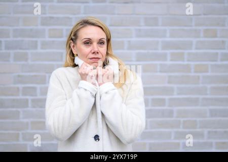 Une femme blonde dans un manteau blanc doux tient son col étroitement, son expression calme et introspective. Banque D'Images