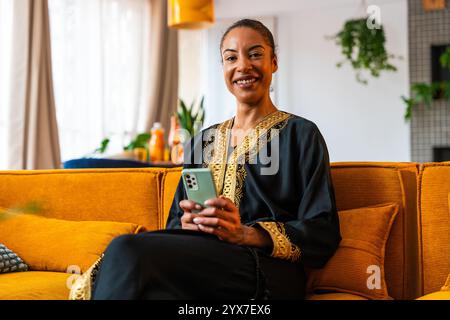 Heureuse belle jeune femme arabe portant traditionnel assis sur un canapé à la maison en utilisant un téléphone intelligent pour magasiner en ligne Banque D'Images