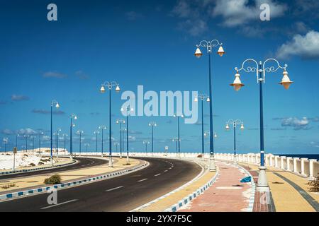 Une promenade côtière pittoresque à Mersa Matruh, en Égypte, avec d'élégants lampadaires bleus, une route sinueuse et un ciel vibrant au-dessus de la Méditerranée S. Banque D'Images