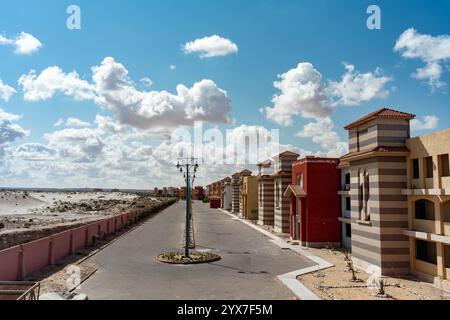 Villas colorées avec des façades rayées et des toits de tuiles bordent une rue calme du Blue Porto Matruh Resort à Mersa Matruh, en Égypte, sous un ciel bleu vif. Banque D'Images