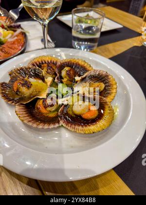 6 coquilles saint-jacques fraîchement cuites dans leurs coquilles sur une assiette à une table à dîner Banque D'Images