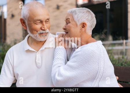 Portrait de belle femme âgée aux cheveux gris touchant doucement l'épaule partenaire partageant un moment chaud et aimant assis sur le banc de la ville le jour d'été. Banque D'Images
