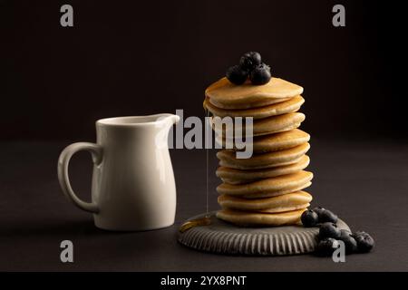 Image de studio sur fond sombre d'une pile de crêpes ou de scones de goutte surmonté de bleuets et d'un pot de sirop. Banque D'Images
