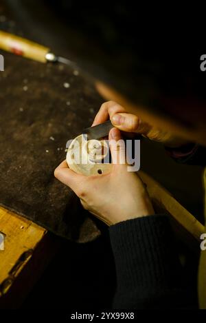 Luthier femelle sculptant méticuleusement un rouleau de violon à l'aide d'un petit ciseau dans son atelier faiblement éclairé, mettant en valeur le savoir-faire complexe impliqué dans cette œuvre Banque D'Images