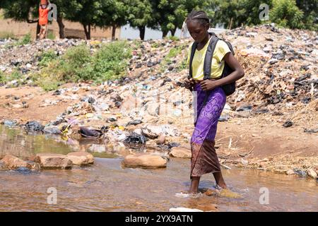 Une jeune écolière africaine observant ses pas alors qu’elle marche dans une petite rivière rocheuse, symbolisant le manque d’infrastructures et de transports en commun Banque D'Images