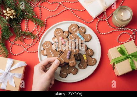 Main tenant des biscuits en pain d'épices sur une assiette blanche décorée de pépites colorées entourées d'un décor de vacances et de cadeaux sur un fond rouge. Top vie Banque D'Images