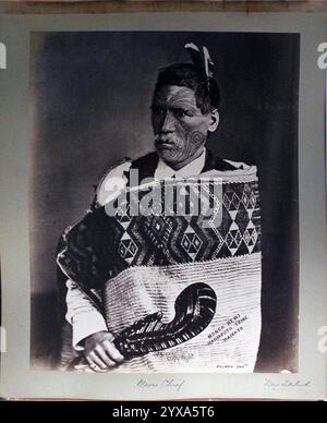 Portrait du chef maori, Monga Rewi Maniapoto, tiré d'une publication de 1942 présentant la collection Victor Millard. Photographié par William Henry Thomas Partington vers 1900, probablement à Wanganui. Rewi pose sur un fond Uni. Il a une fine barbe blanche, des cheveux courts et blancs décorés de plumes et de tatouages faciaux étendus. Il porte un manteau rigide orné de motifs géométriques autour de ses épaules avec une chemise européenne et un gilet en dessous. Il tient une courte pagaie sculptée dans ses mains, peut-être un symbole d'autorité. Banque D'Images