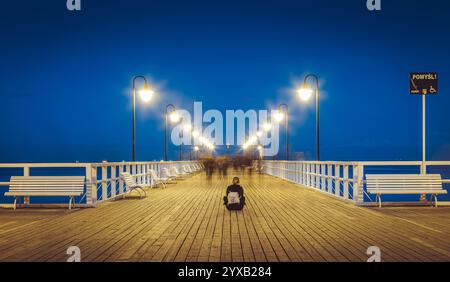 Jeune femme assise sur une jetée en bois à gdynia orlowo, sopot, pologne, trempant dans la vue nocturne imprenable sur la mer baltique, illuminée par de douces lumières Banque D'Images