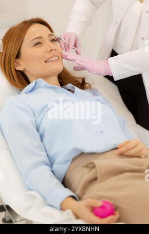 Le patient se détend sur un lit de traitement confortable tandis qu'un praticien qualifié applique un produit de remplissage dermique pour améliorer les traits du visage dans une clinique tranquille envi Banque D'Images