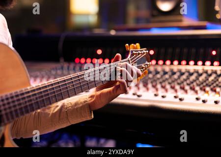 Chanteur qualifié jouant sur sa guitare électro-acoustique dans la salle de contrôle, enregistrant de nouveaux sons et mixant avec la console audio. Musicien et technicien composant une chanson à succès pour un album. Gros plan. Banque D'Images