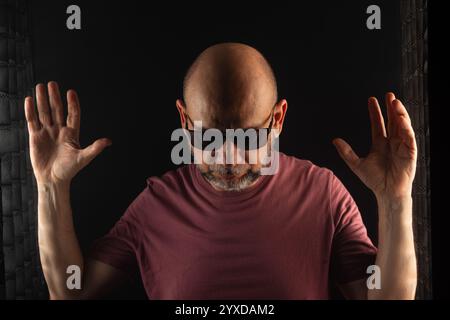 Homme chauve et barbu portant des lunettes de soleil avec les mains levées. Isolé sur fond fumé foncé. Personne sud-américaine. Banque D'Images