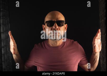 Homme chauve et barbu portant des lunettes de soleil avec les mains levées. Isolé sur fond fumé foncé. Personne sud-américaine. Banque D'Images