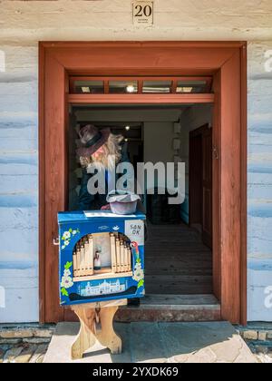 Sanok, Pologne - 4 mai 2024. Un musicien de rue barbu en chapeau et manteau se tient à la porte d'une maison en bois, jouant d'un orgue à baril peint à la main. Banque D'Images