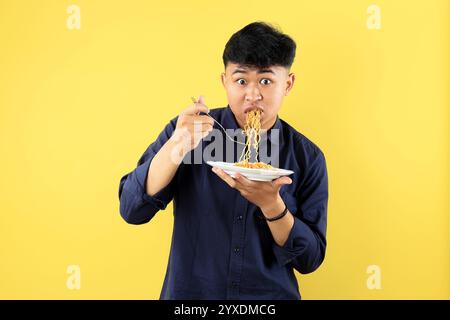 Jeune garçon indonésien grands yeux ouverts, WOS geste tout en mangeant Indomie Mie Goreng sur plaque blanche, espace de copie sur fond jaune Banque D'Images