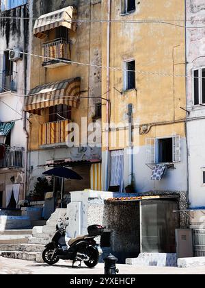 Une charmante scène urbaine rustique avec un bâtiment jaune altéré avec des balcons et des auvents rayés, accompagné d'un scooter stationné. Aventure et Banque D'Images